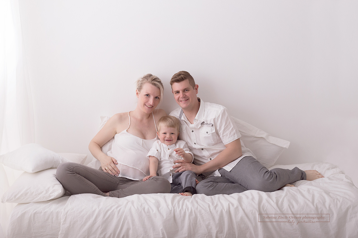glücklich lachendes Familienfoto zu Dritt mit Babybauch auf dem Studiobett