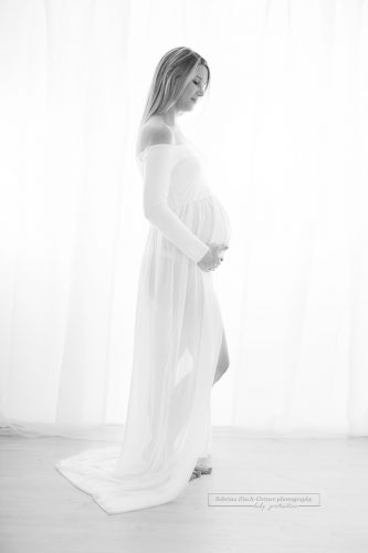 elegantes schwarzweiss Foto in Hochformat einer bald Mama, welche in einem weißen langärmligen Schwangerschaftskleid gehüllt ist.