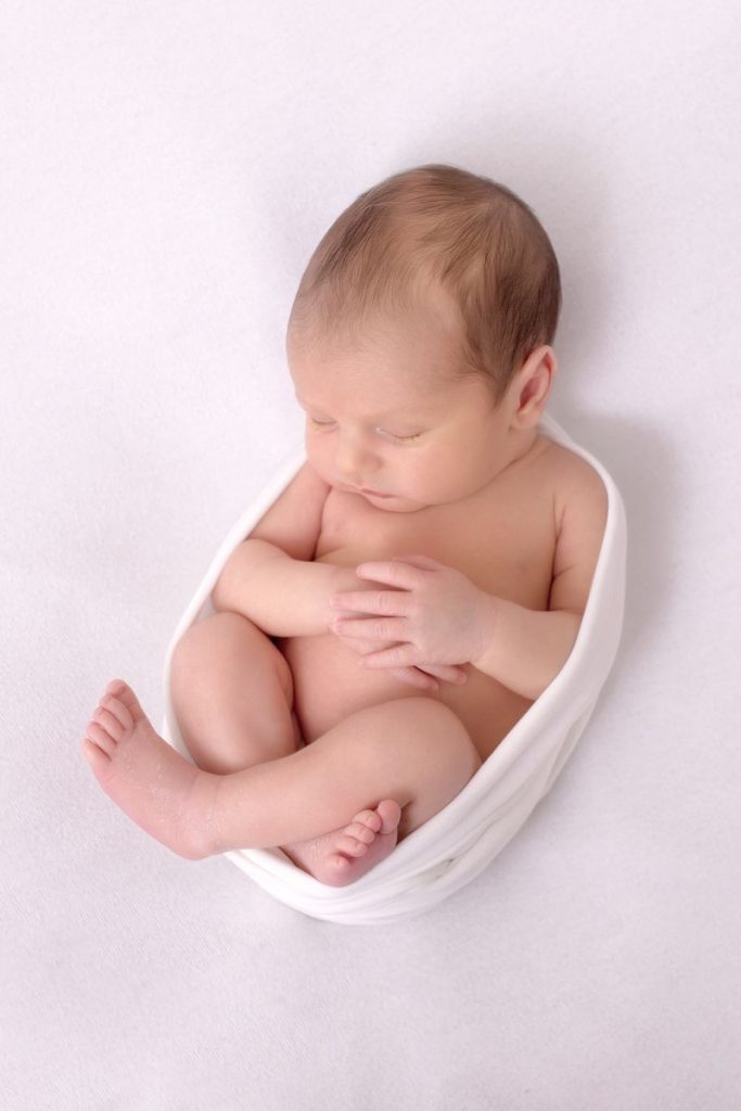 FAQ Neugeborenen Shooting Bild zeigt ein neugeborenes Baby eingewickelt in einen weißen Stoffwrap gemütlich schlafend