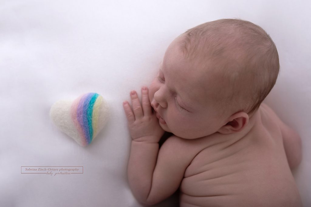 Filzherz mit kleinem Regenbogen liegt neben kleinem Neugeborenen