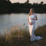 Schwangere im weißen Bellykleid vor mit Abendlicht getränktem Wasser