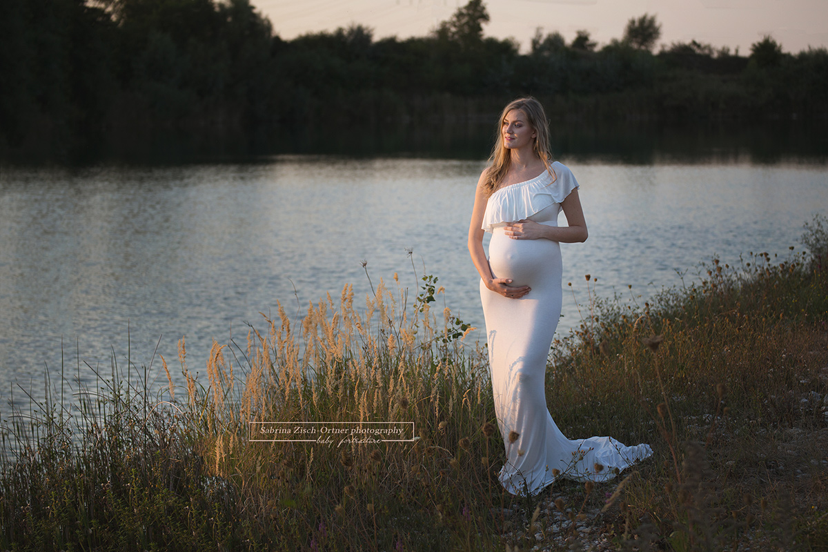 Schwangere im weißen Bellykleid vor mit Abendlicht getränktem Wasser