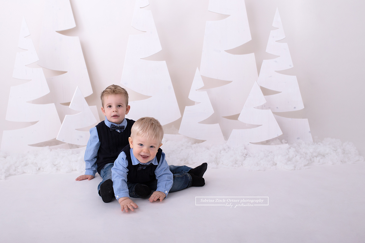 zwei Brüder sitzend und lachend vor den Weihnachtsbäumen im Alter von 1 und 3