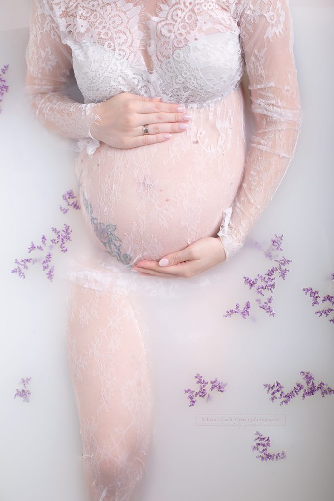Babybauch in einem Bad aus Milch in Spitzenkleid und Lavendelblueten