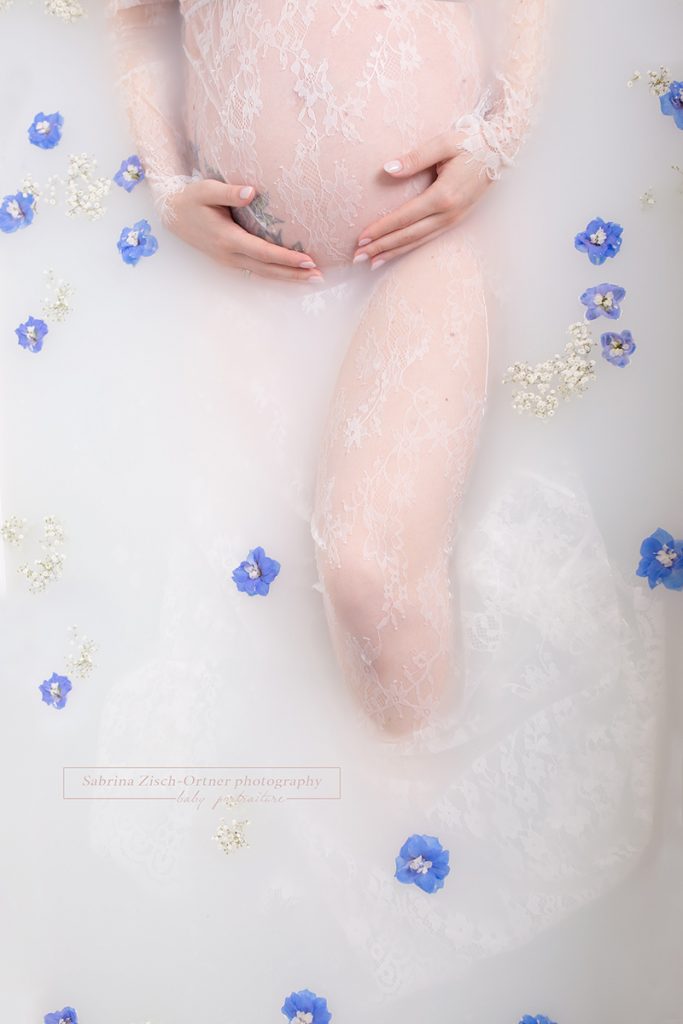 Babybauch umhuellt vom weissen Spitzenkleid in der Badewanne mit Milch und blauen Blumen