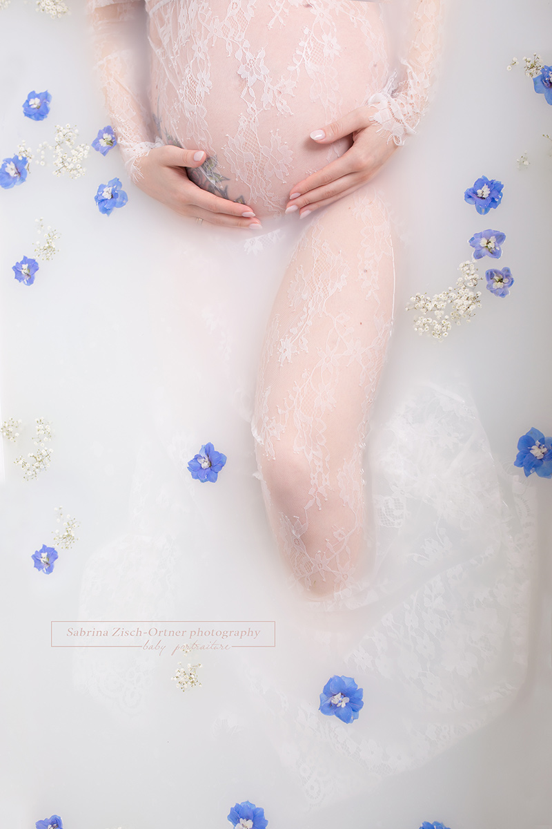 Babybauch umhüllt vom weißen Spitzenkleid in der Badewanne mit Milch und blauen Blumen