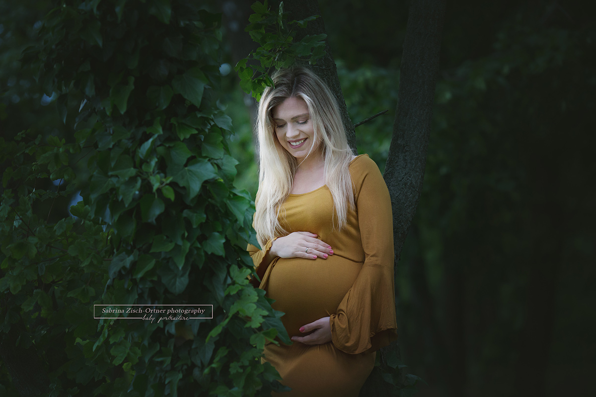 Bald Mama bei ihrem Fotoshooting mit Babybauch im gelben Kleid inmitten vom dunkelgrünen Efeu