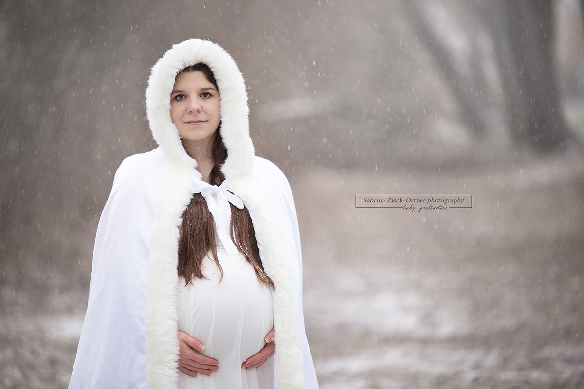 Bald Mama umhüllt ihren Babybauch mit weißen Wintermantel bei ihrem Outdoorshooting