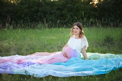 Schwangere sitzt in Wiese umhüllt von einem meterlangen Regenbogenrock