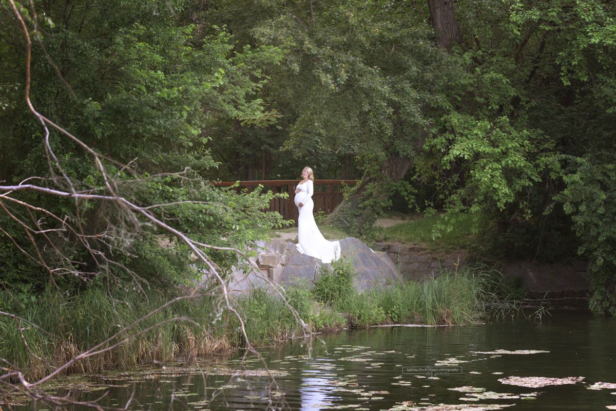 Babybauch Outdoorshooting im weißen Kleid von grüner Natur und Wasser umgeben