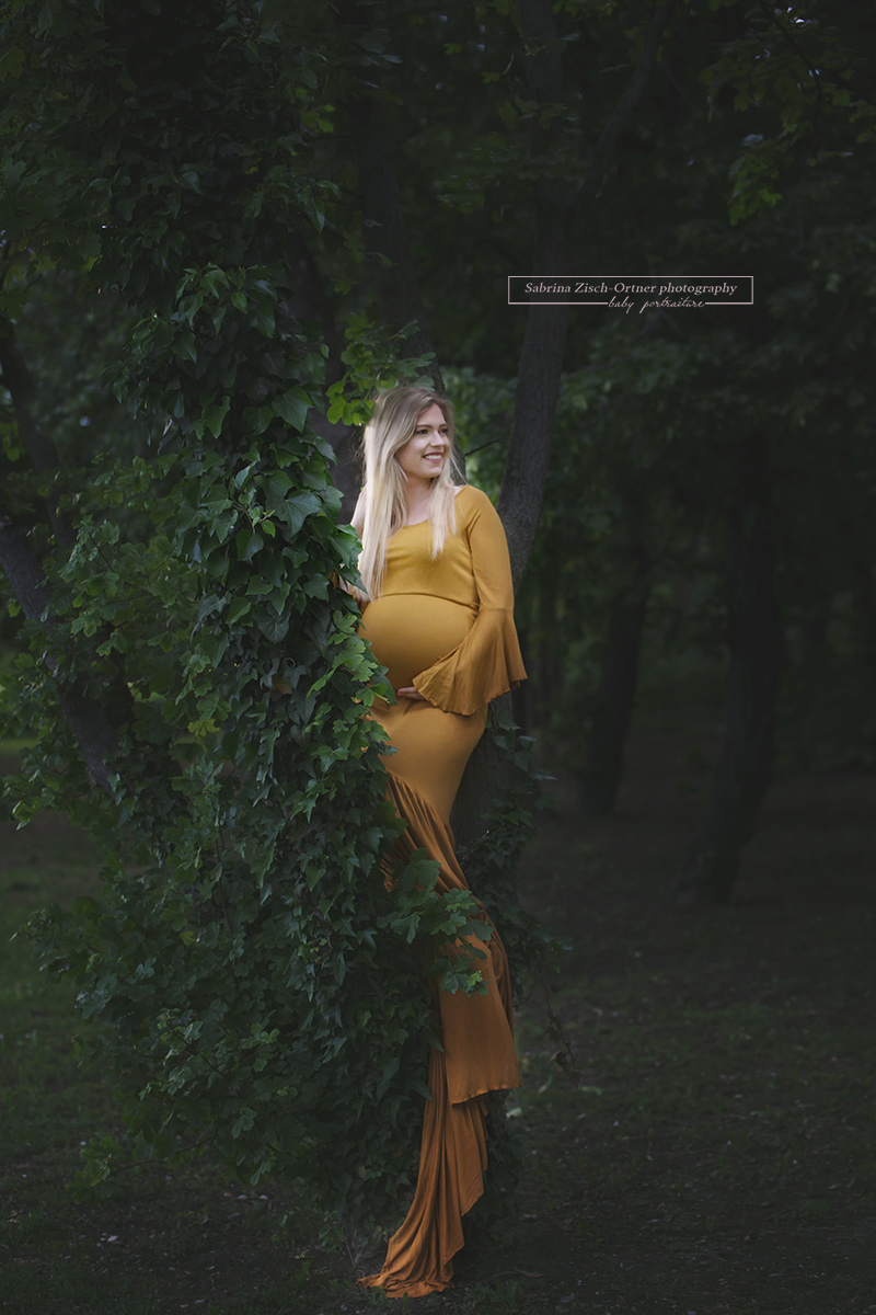 Ein Baum mit sattem grünen Efeu bedeckt und eine Schwangere im gelben Kleid mit meterlangen Schleppe