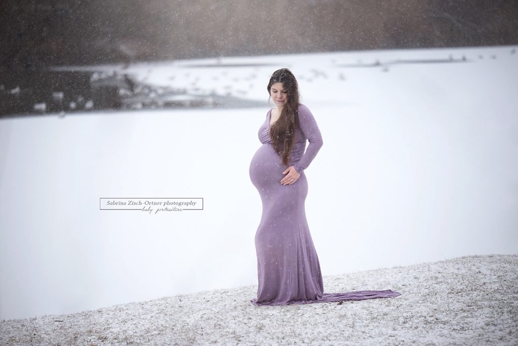 Schwangere im lila Babybauchkleid vor zugefrorenen und verschneiten See