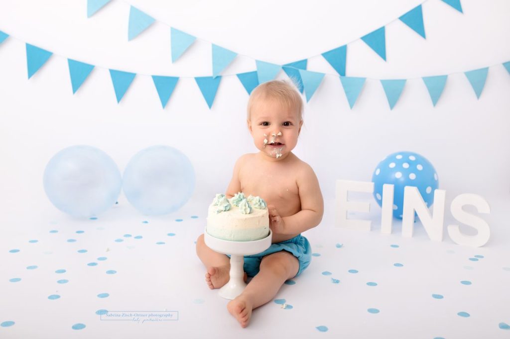 lachender Bub vor seiner Geburtstagstorte mit Wimpelketten und Luftballons in blau