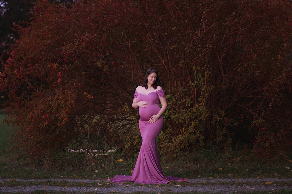 Schwangere im figurbetonten rosa Babybauchkleid vor rotem Busch