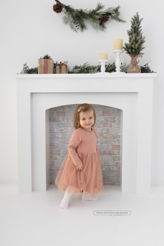 kleines grinsendes stehendes Mädchen in Rosa vor weihnachtlich weißem Kamin
