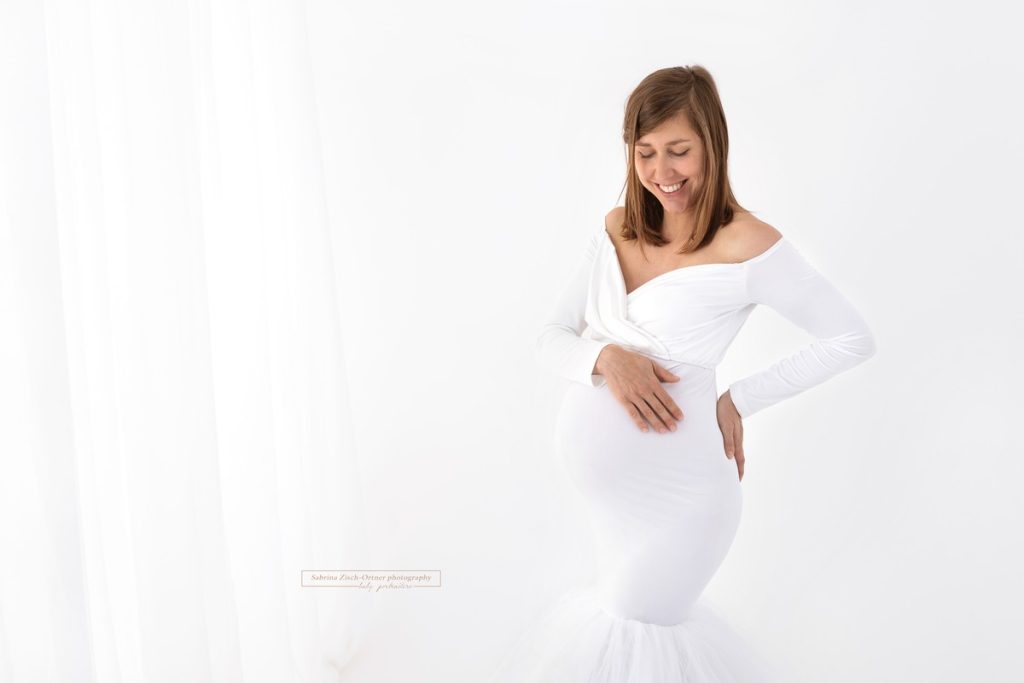 lachende bald Mama in weißem Schwangerschaftskleid bei Fotoshooting